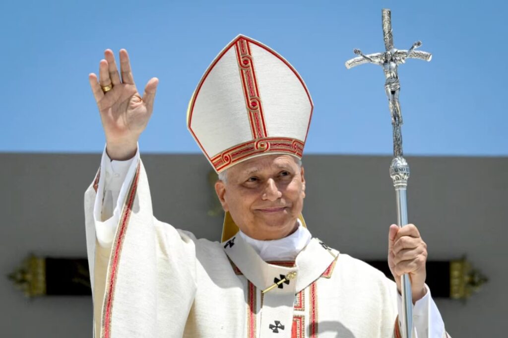 El papa León XIV, con mitra y vestiduras blancas, saluda con la mano en alto y sostiene el báculo durante una misa al aire libre.