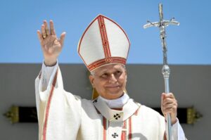 El papa León XIV, con mitra y vestiduras blancas, saluda con la mano en alto y sostiene el báculo durante una misa al aire libre.